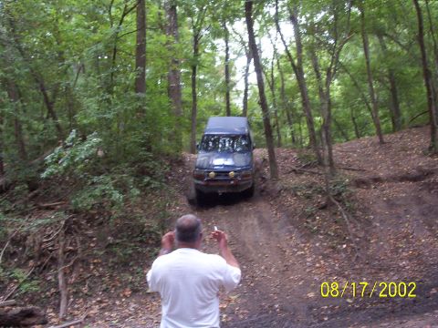 1991 Ford Bronco - A blue SUV navigates a dirt path through a dense forest with a man standing in the foreground.