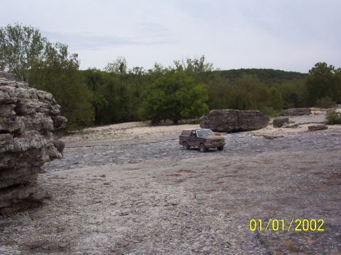 1991 Ford Bronco - A brown Ford Bronco is parked on a rocky surface with trees and hills in the distance.