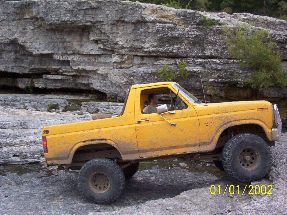Yellow Ford Bronco with large tires driving over rocks, driver visible. Water and rocky terrain