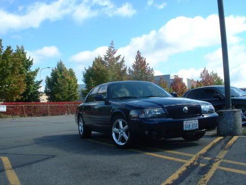 1999 Ford Crown Victoria - A dark blue Mercury Grand Marquis is parked in a lot on a cloudy day, with trees in the background.