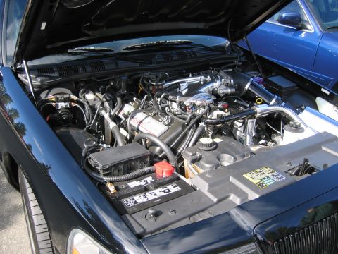 1999 Ford Crown Victoria - The engine bay of a black car is shown, with another blue vehicle visible behind it.
