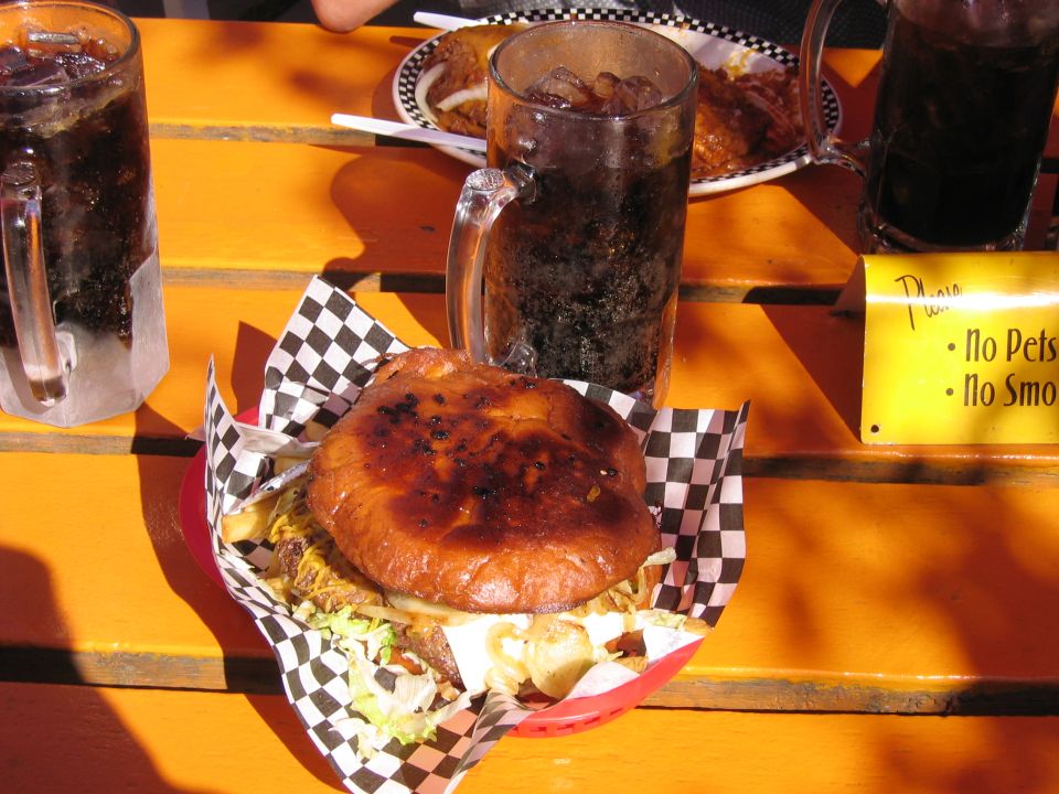 Burger with fries, two mugs of soda, and a plate of food on an orange table. A yellow sign says 'No Pets, No Smoke'