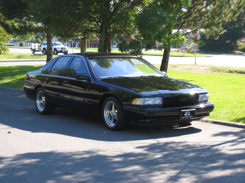 Black Chevrolet sedan with aftermarket wheels parked on asphalt under trees