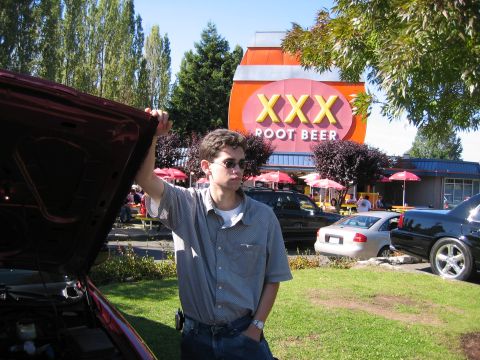 1999 Ford Crown Victoria - A man stands by the open hood of a car, with a distinctive XXX Root Beer stand in the background and other vehicles parked nearby.