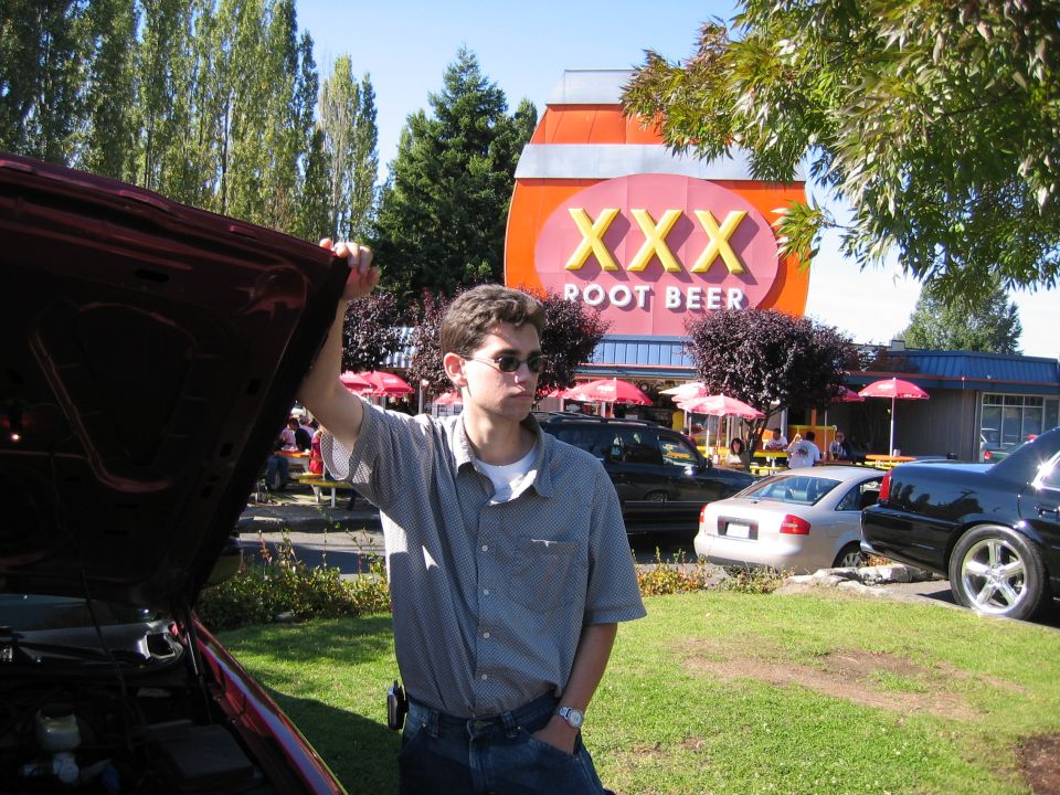 Man by open car hood, XXX Root Beer sign behind. Several cars visible
