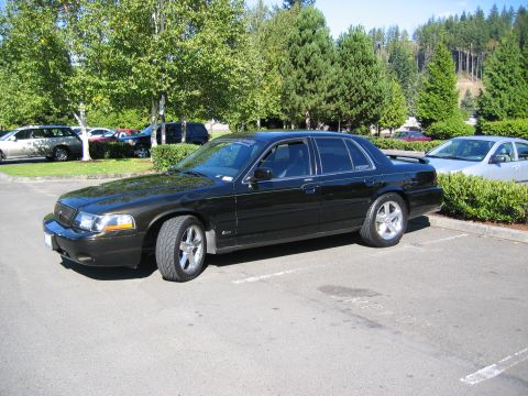 1999 Ford Crown Victoria - A black sedan is parked in a lot with other vehicles and trees in the background.