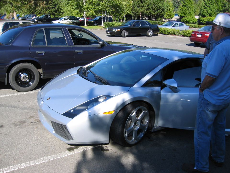 Silver sports car with door open, parked next to a dark sedan. Man in blue shirt and jeans stands by the sports car