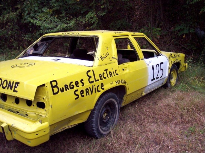 Yellow gutted demolition-derby sedan number 125 lettered Burges Electric Service in a field