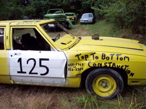 1987 Mercury Grand Marquis - A yellow demolition derby car is parked in a grassy area with other vehicles in the background.