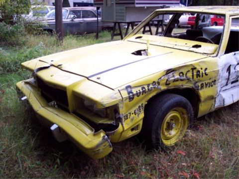 1987 Mercury Grand Marquis - A yellow sedan, likely prepared for a demo derby, shows significant front-end damage and custom lettering.