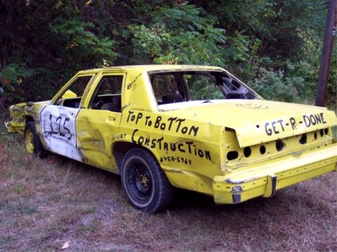 1987 Mercury Grand Marquis - This heavily modified yellow sedan, likely prepared for demolition derby, displays custom text and significant body damage.