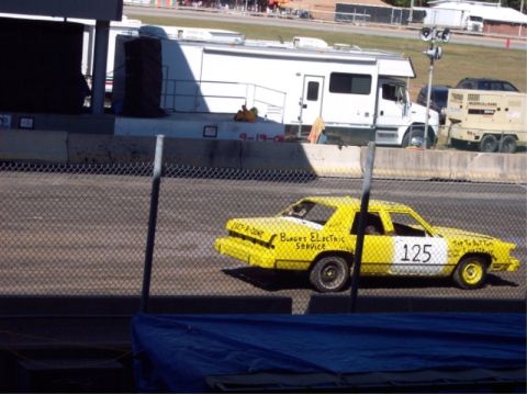 1987 Mercury Grand Marquis - A yellow sedan, marked with '125' and 'Burges Electric Service', is seen on a track, likely for a demolition derby event.