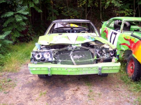 1987 Mercury Grand Marquis - Two heavily damaged green sedans sit side-by-side in a grassy area, suggesting they were used in a demolition derby.