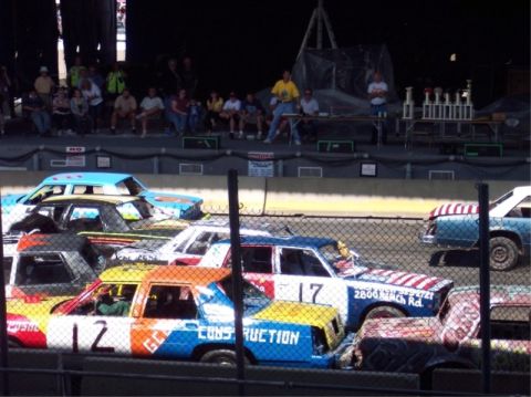 1987 Mercury Grand Marquis - A group of cars are gathered on a track, likely for a demolition derby event, with spectators in the background.