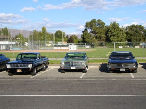 1967 Ford Ranchero - Three classic pickup trucks, including a black 1967 Ford Ranchero, are parked in a lot with a baseball field in the background.