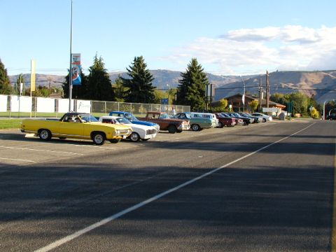 1967 Ford Ranchero - A line of classic cars are parked in a lot, led by a yellow 1970-1971 Ford Ranchero.