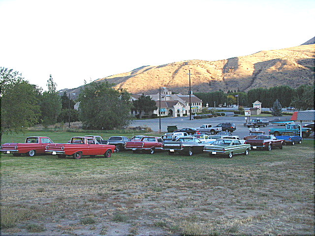 Rows of 1960s Ford cars and Rancheros parked on grass at a club gathering with hills behind