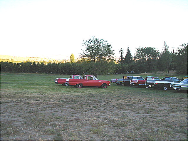 Red early-1960s Ford sedan in profile with a row of classic cars in a field