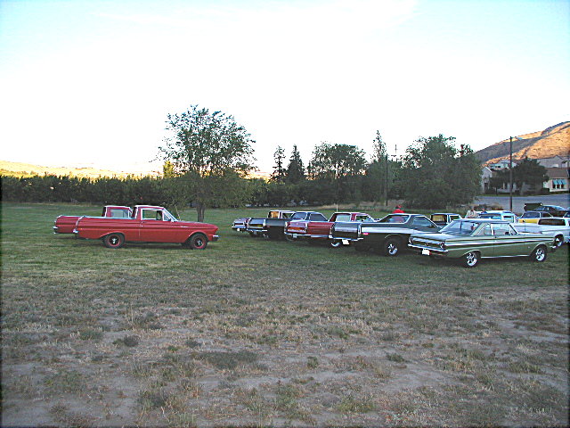 Red 1960s Ford Ranchero in profile leading a row of classic cars on grass at dusk