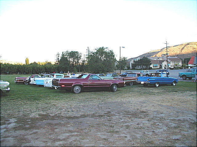 Classic cars lined up on grass at dusk car show, maroon car-based pickup in center