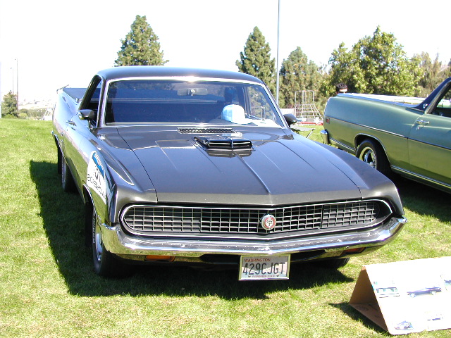 Front view of a dark gray Ford Ranchero with a hood scoop and a Washington license plate. Another green car is parked next to it