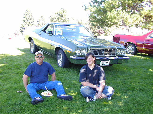 Two people sitting on grass in front of a green 1970s Ford Ranchero at a car show