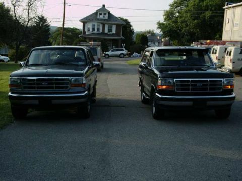 1992 Ford Bronco - Two black Ford Broncos are parked on a street in front of a house, with vans visible in the background.