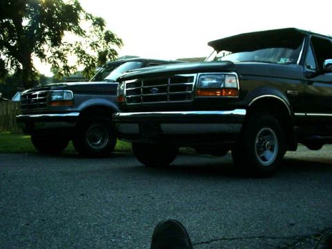 1992 Ford Bronco - Two Ford Broncos are parked on asphalt, with trees and a fence in the background.