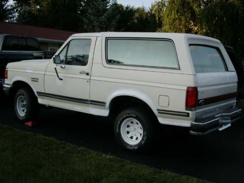 1988 Ford Bronco - The rear three-quarter view of a white SUV is shown parked on a dark surface next to a dark pickup truck.