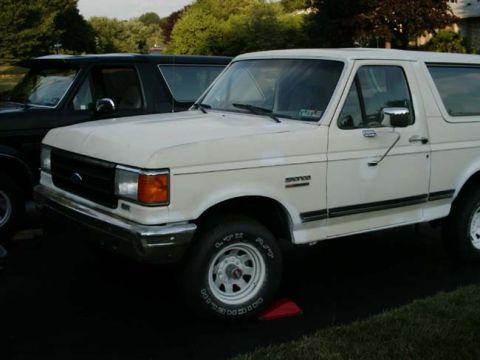 1988 Ford Bronco - A white Ford Bronco is parked next to a black SUV, with trees and a building in the background.