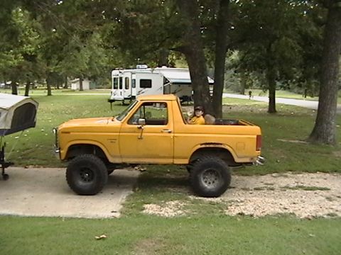 1986 Ford Bronco - A lifted yellow Bronco sits parked on a gravel patch next to a campsite, with a camper in the background.