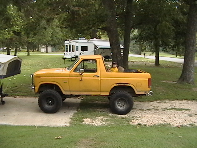 Yellow lifted Bronco parked on gravel, with a person in the back seat. Camper trailer in background