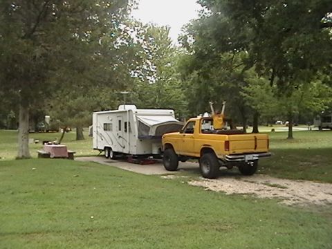 1986 Ford Bronco - A yellow pickup truck tows a pop-up camper through a wooded area, with a person in the truck bed raising their arms.