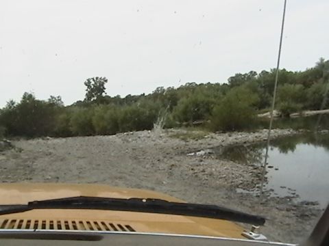 1986 Ford Bronco - Driving a vehicle through shallow water near a wooded riverbank.