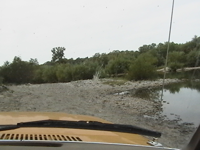 Yellow vehicle hood and windshield wipers in foreground, driving through shallow water with trees and river in background