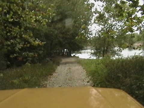 1986 Ford Bronco - View from inside a yellow vehicle driving down a dirt path towards a body of water surrounded by trees.