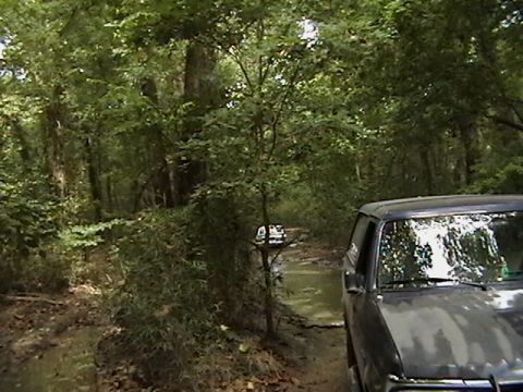 1986 Ford Bronco - A dark SUV navigates a muddy path through a wooded area, with another vehicle visible in the distance.