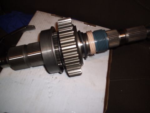 1985 Ford Bronco - A transfer case or transmission output shaft laid on cardboard, carrying a splined hub, a gear, a bearing race and a teal-green threaded/co…