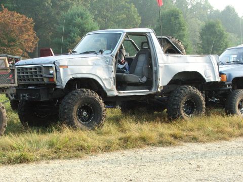 1996 Ford Bronco - This heavily modified SUV, likely from the early 1980s, sits parked on a dirt road next to another vehicle.