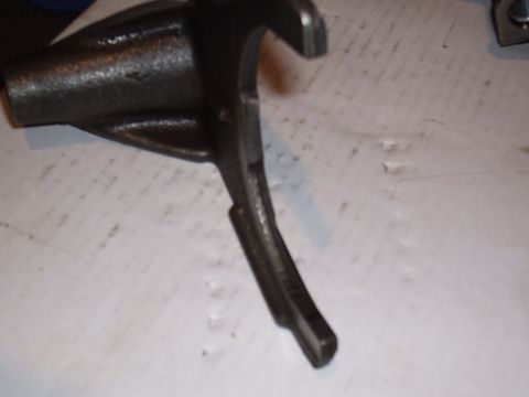 1985 Ford Bronco - Extreme close-up of a cast-steel transmission shift fork lying on a white paper-covered workbench, with its cylindrical shaft boss at left…
