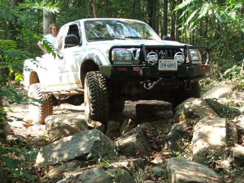 1996 Ford Bronco - Toyota Tacoma navigating a challenging rocky off-road course in a wooded area.