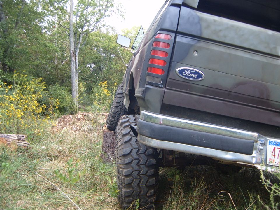 Dark green Ford Bronco with camouflage pattern, lifted on large tires, parked on a log, rear view