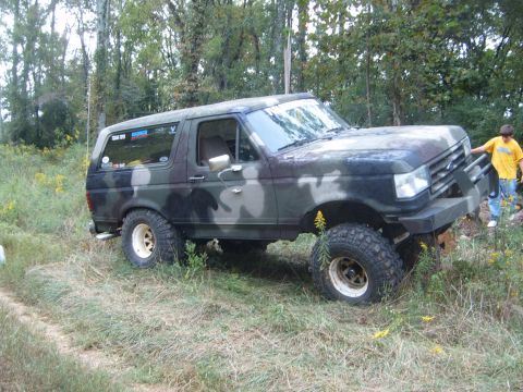 1988 Ford Bronco - Camouflaged 1988 Ford Bronco with off-road modifications sits in a field with trees in the background.
