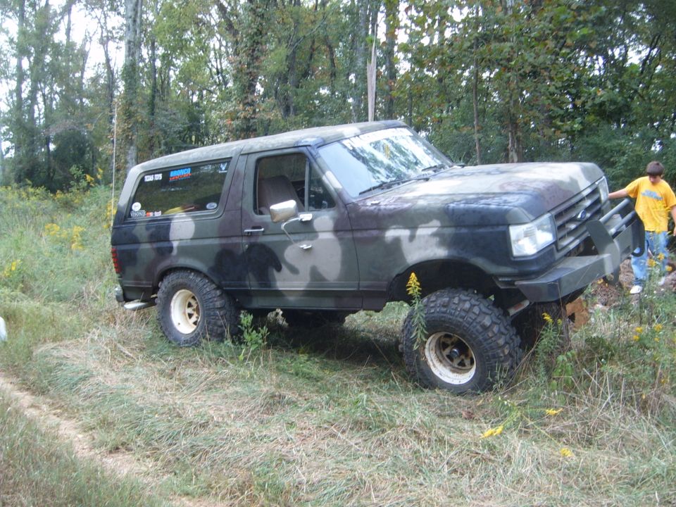 Camouflage-painted lifted Ford Bronco on mud tires parked on a grassy trail