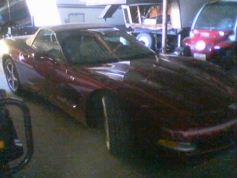 1990 Ford Bronco - dark maroon Chevrolet Corvette coupe sits parked in a cluttered garage next to a red SUV and other equipment.
