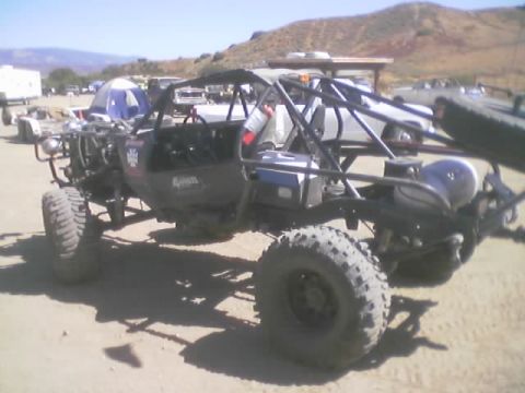 1990 Ford Bronco - Rear view of a silver and black tube-frame rock buggy with roll cage and exposed fuel cell parked at a dusty desert gathering.