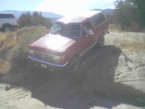 1990 Ford Bronco - red Ford Bronco with a white roof eases down a rutted sandy trail in the desert as the driver waves from the open window.