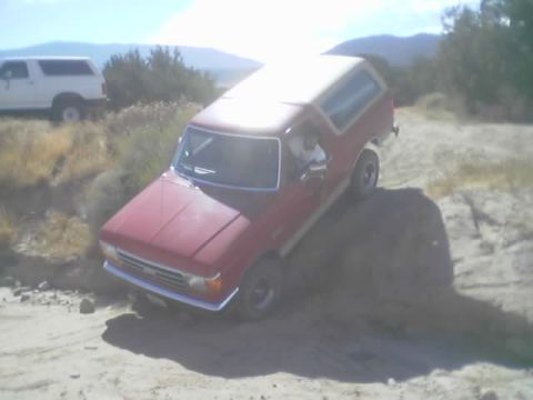 1990 Ford Bronco - red Ford Bronco with a white roof noses down into a sandy desert rut, kicking up dirt, with a passenger visible inside.