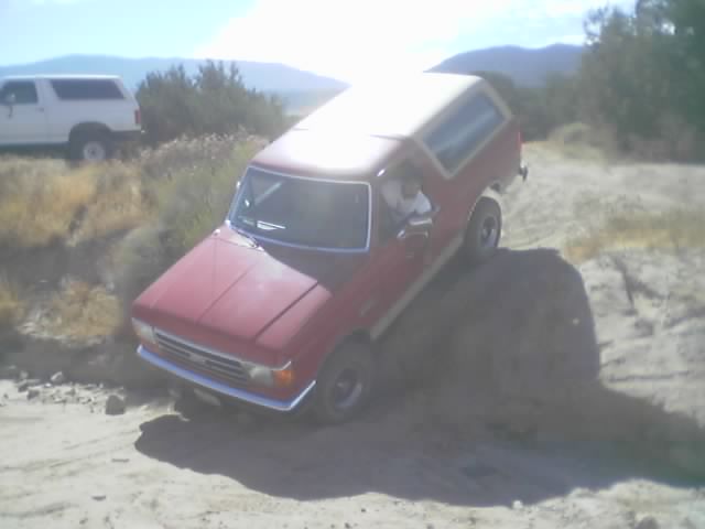 Red Ford Bronco with white top dropping into a sandy desert rut, driver inside
