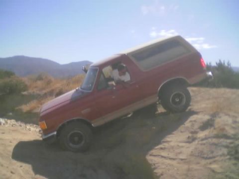 1990 Ford Bronco - red Ford Bronco with tan lower bodywork sits tilted on a sandy desert slope under a clear blue sky.